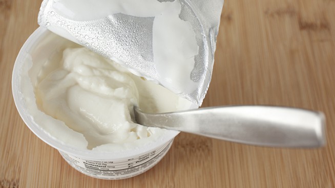 A small container of greek yogurt with the spoon in it and the foil covering pulled back.